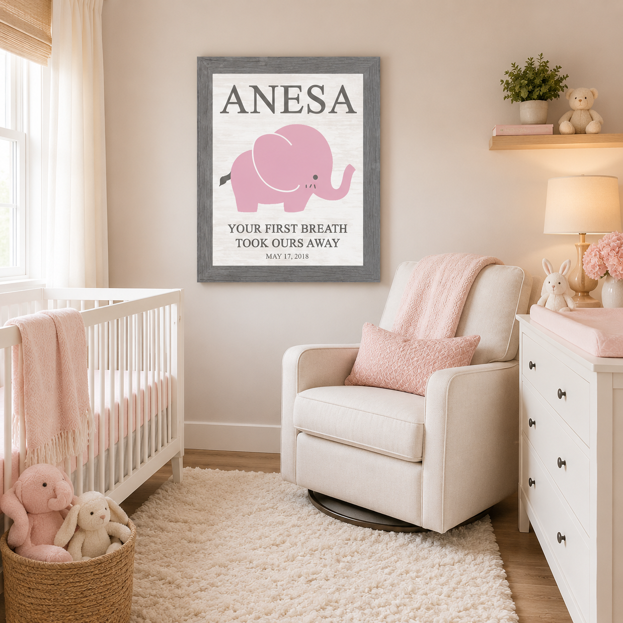 Gray framed personalized baby name nursery wall art PER149 displayed large in soft pink girl nursery above glider chair beside white crib — pink elephant with bold name ANESA at top and Your First Breath Took Ours Away with birth date May 17 2018 on soft white background — stuffed bunnies changing table and white dresser in room