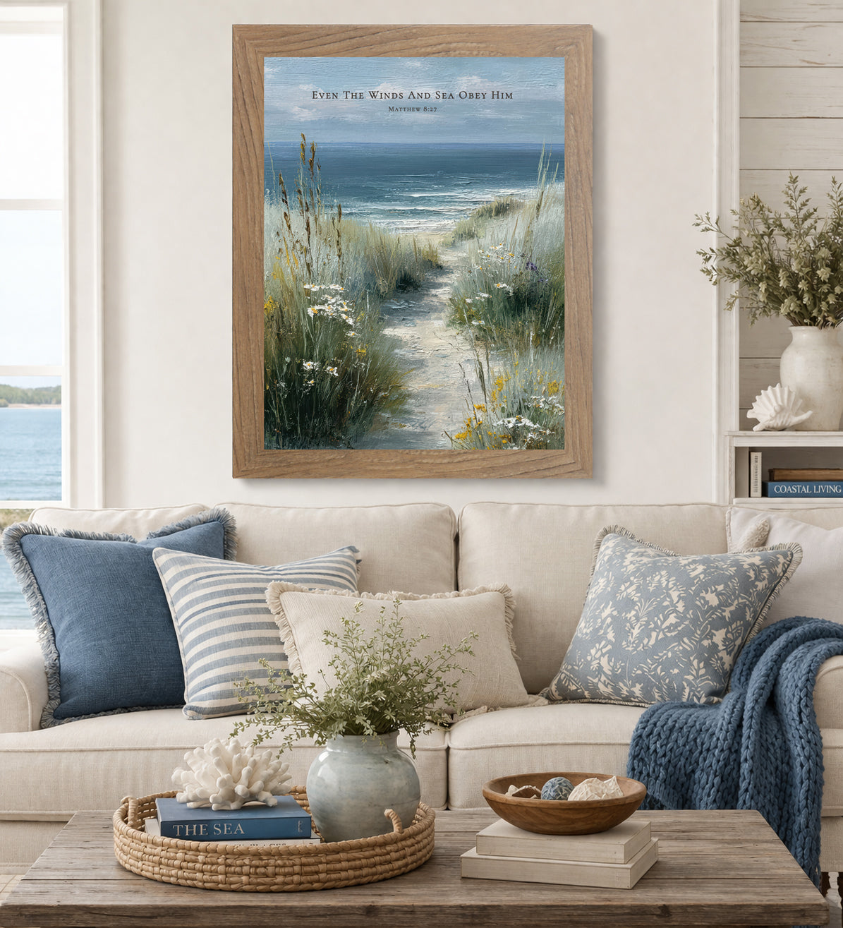 Farmtable framed Even The Winds And Sea Obey Him Matthew 8:27 coastal portrait wall art displayed above a cream sofa in a coastal living room with blue and striped pillows blue knit throw and seashell decor with water visible through the window