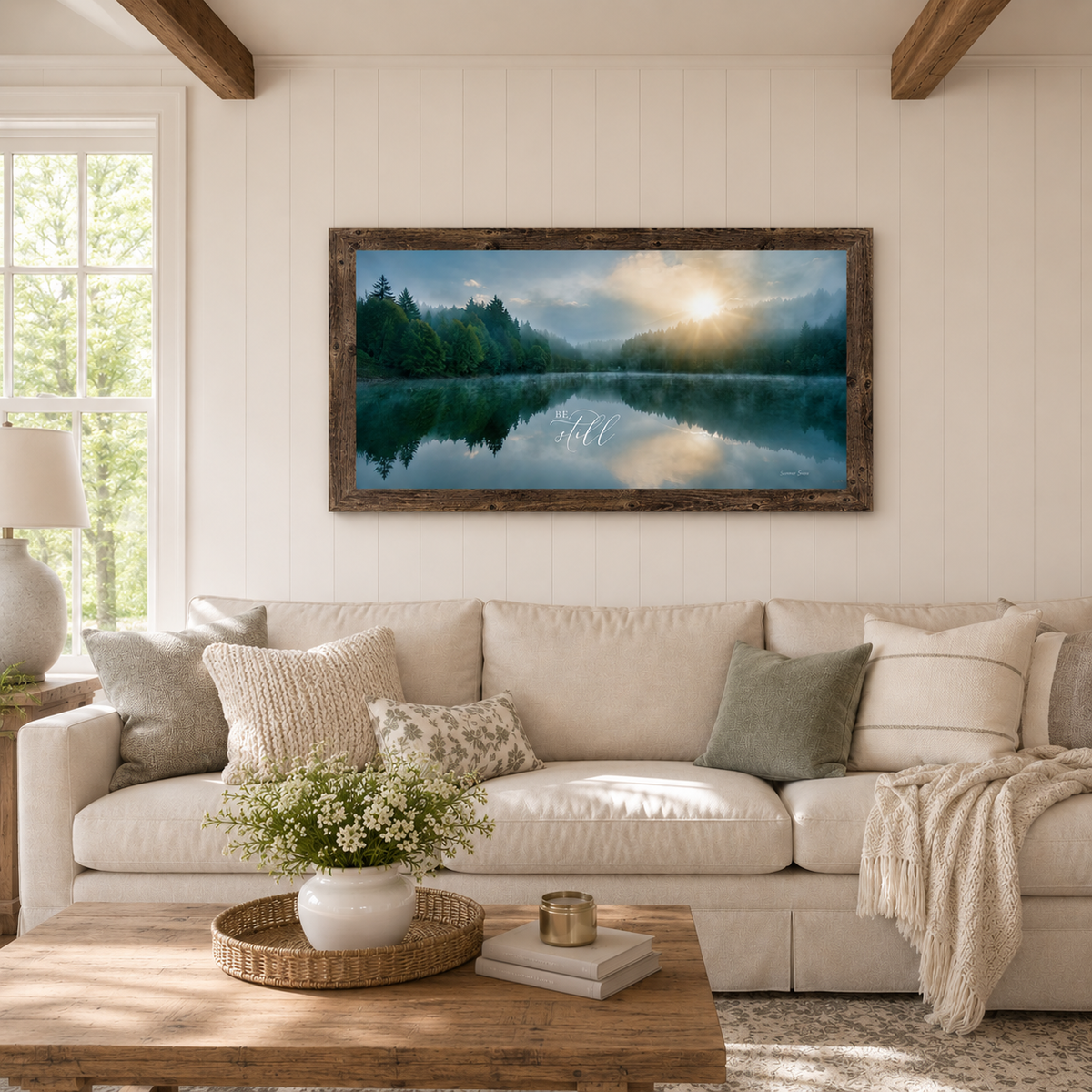 Natural brown farmtable framed Be Still wide landscape wall art SB765 displayed large above cream linen sofa in bright farmhouse living room with shiplap walls wood beam ceiling and white floral centerpiece — misty lake sunrise with pine forest reflection and golden sun breaking through clouds with elegant Be Still script typography