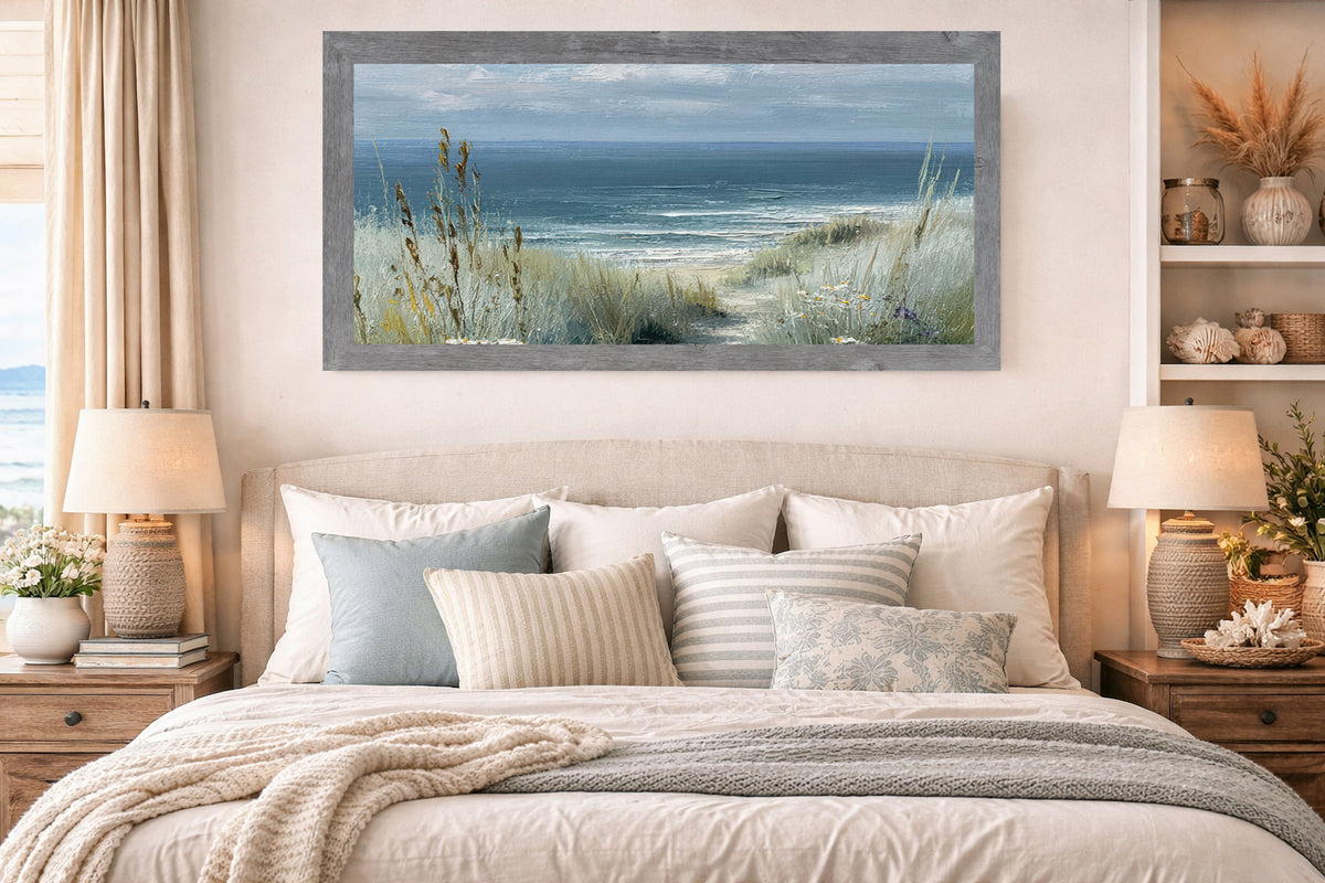 Gray framed coastal path wide landscape wall art displayed above a bed in a coastal bedroom with neutral linen headboard blue and striped pillows woven lamps seashells on shelves and pampas grass