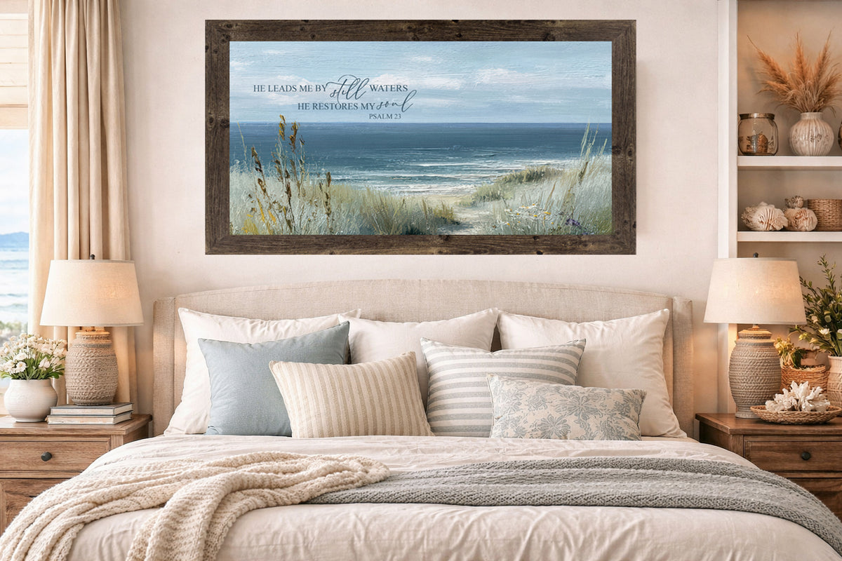 Natural brown framed He Leads Me By Still Waters Psalm 23 coastal wide landscape wall art displayed above a bed in a coastal bedroom with neutral linen headboard blue and striped pillows woven lamps and seashell decor on shelves