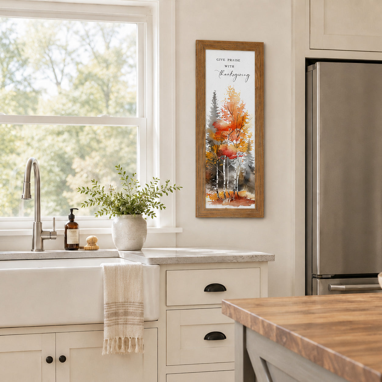 Farmtable framed Give Praise With Thanksgiving narrow portrait fall wall art SN785 displayed in bright farmhouse kitchen beside window between sink and refrigerator — vivid watercolor autumn orange red and birch forest trees with elegant mixed script typography — butcher block island farmhouse sink and greenery in white vase