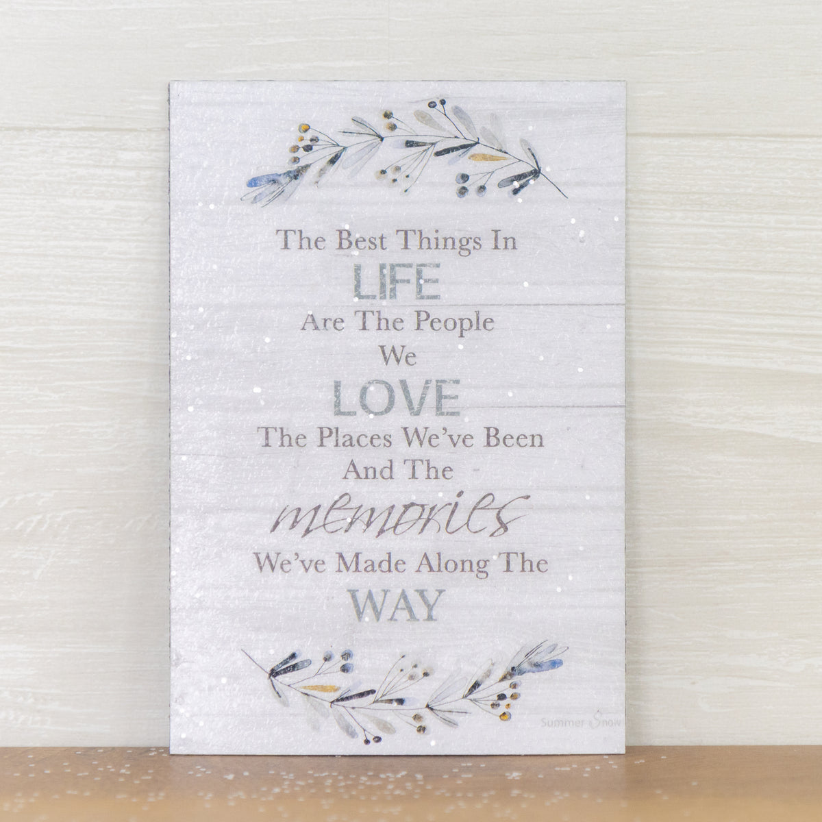 Inspirational quote magnet featuring The Best Things in Life saying on gray slatted wood background with floral accents, neutral vertical decor with subtle glitter finish